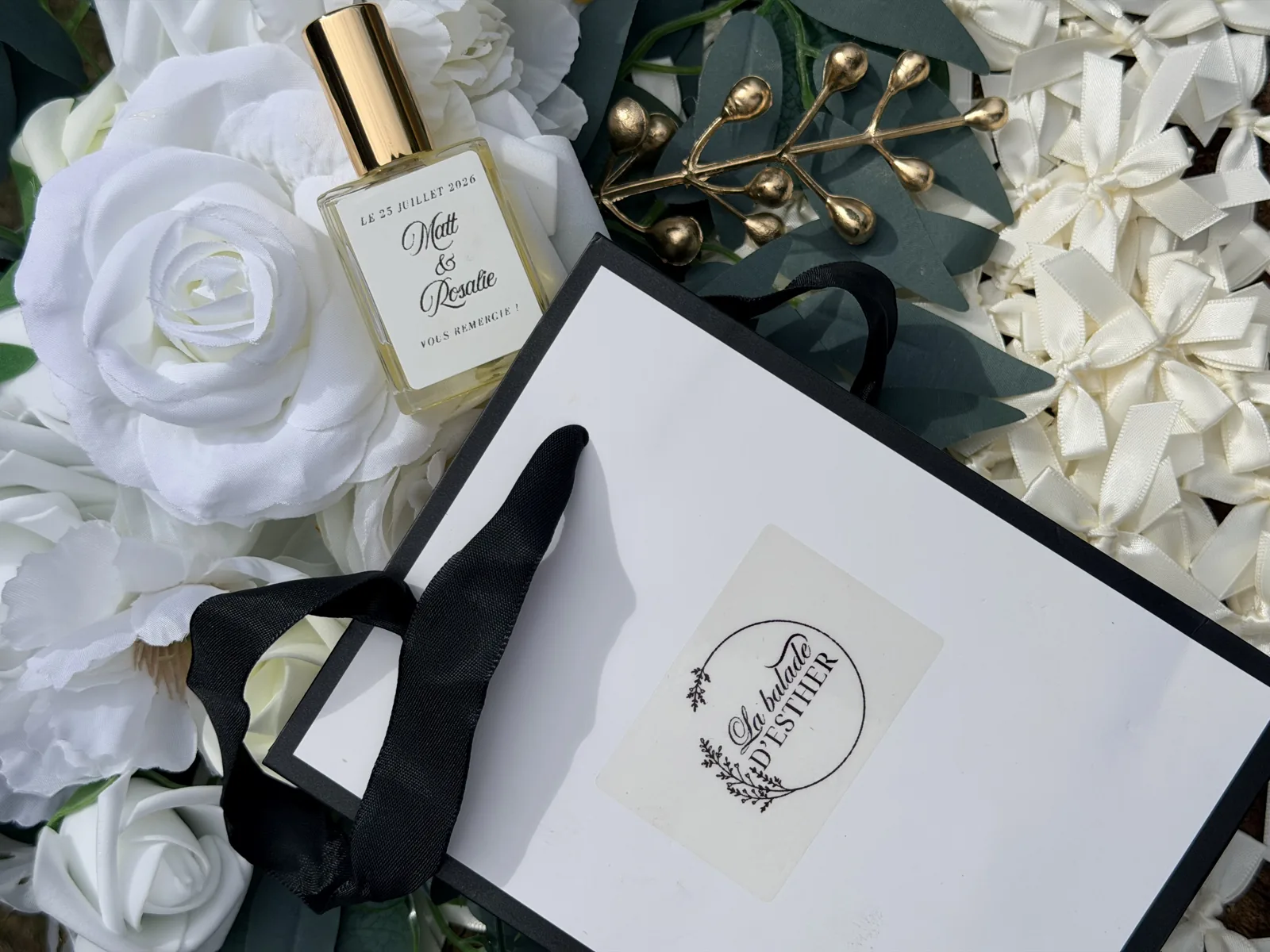 Flacon étiqueté Matt et Rosalie, 25 juillet 2026, posé sur des roses blanches à côté d'un sac cadeau